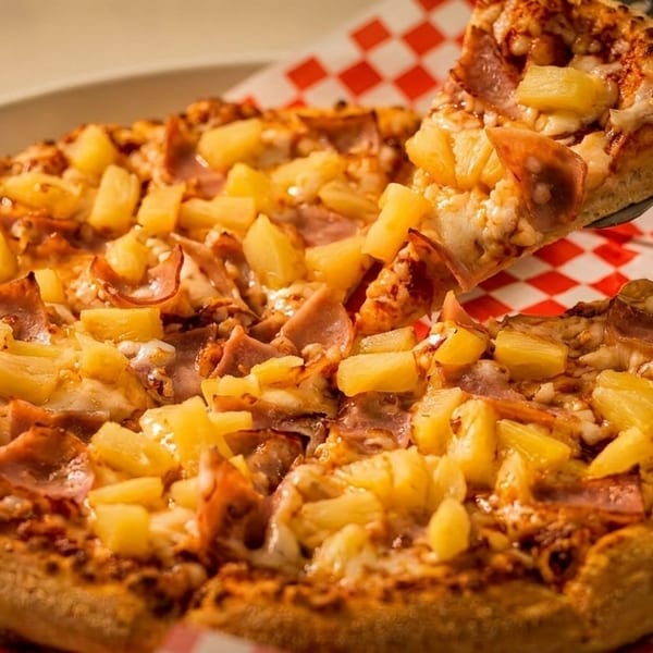 A slice of Hawaiian pizza with ham and pineapple being lifted from a whole pizza on a red checkered paper.