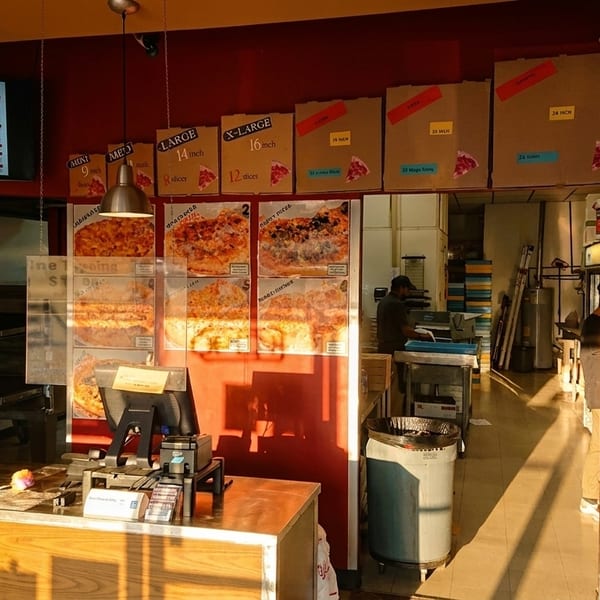 A pizza shop interior with a counter, menu, pizza boxes, and a person standing.